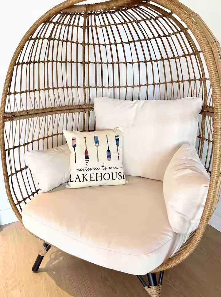Wicker, egg-shaped chair with cream cushions in a bright room, featuring a decorative pillow that says 'Welcome to our LAKEHOUSE' with paintbrush icons.
